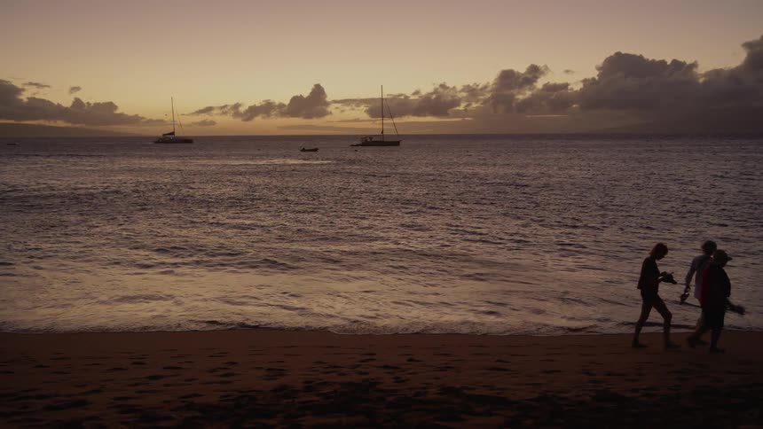 Silhouetted people walk across beach near surf as sailboats float against a red horizon