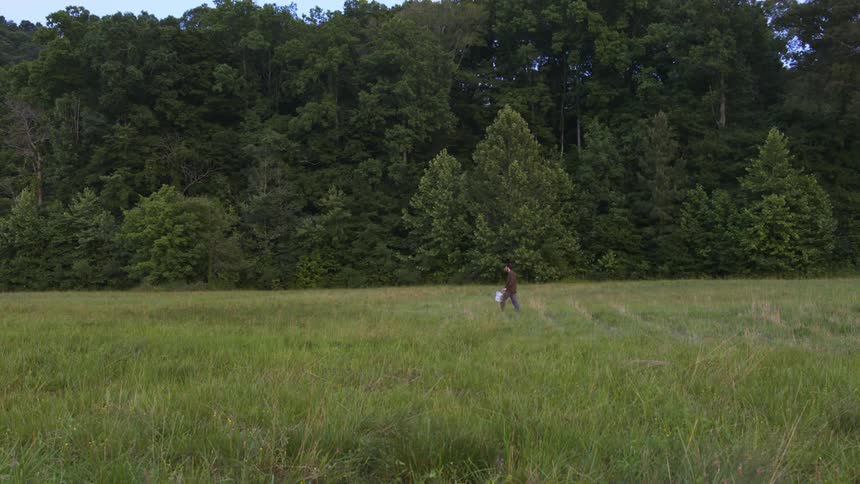 Young man carrying a bucket on a grass field near a forest