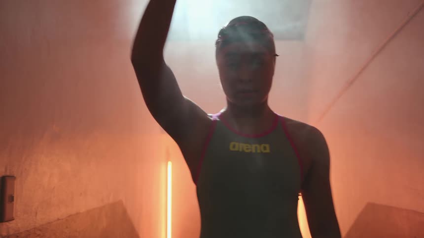 Female swimmer swings arms while walking down smoky corridor lit by neon lights as she preps for a competition