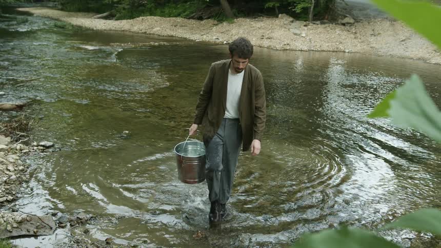 Man with beard brown shirt and blue pants takes bucket of water from the river in the stream
