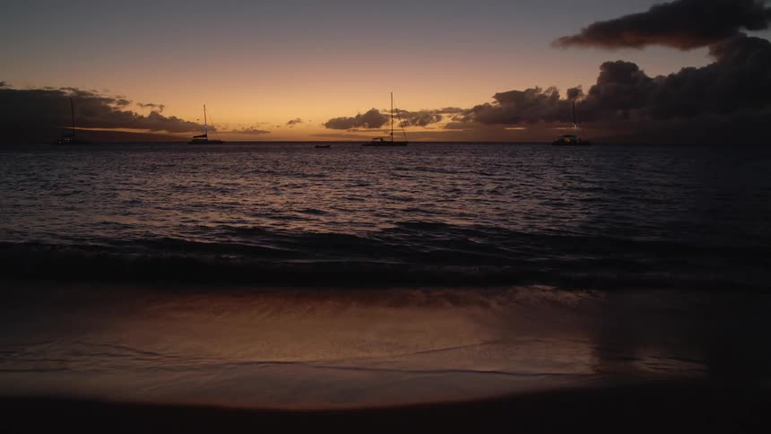 Sailboats with their sails down sit near horizon under a red sky as surf crashes onto beach sand