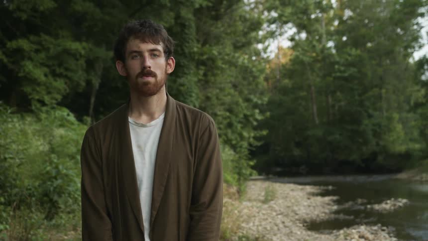 Man with red beard brown and white shirt looks at camera standing next to stream in the forest