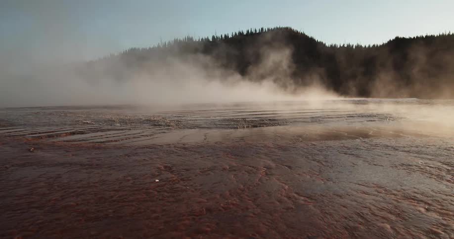 Vapor rises off hot water flowing across ground next to forest covered mountain under a clear blue sky