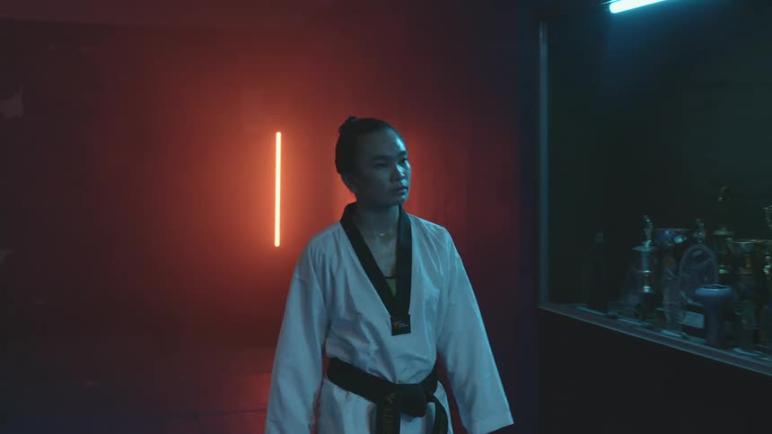 Woman wearing martial arts uniform walks past trophy case in hall lit by red neon light