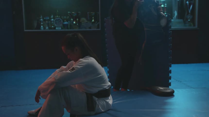 Young woman wearing karate uniform sitting on the floor while someone picks up gear from the floor