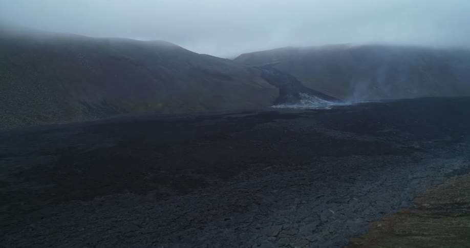 Smoke rises from cooling lava covered ground between mountain and field on an overcast day
