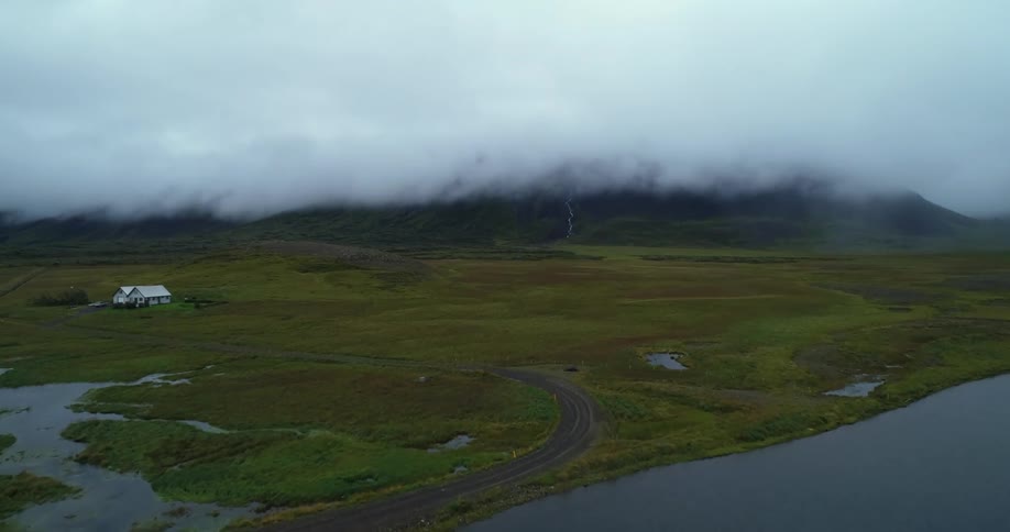 Lone house sits near road curving around water with cloud covered mountain on horizon