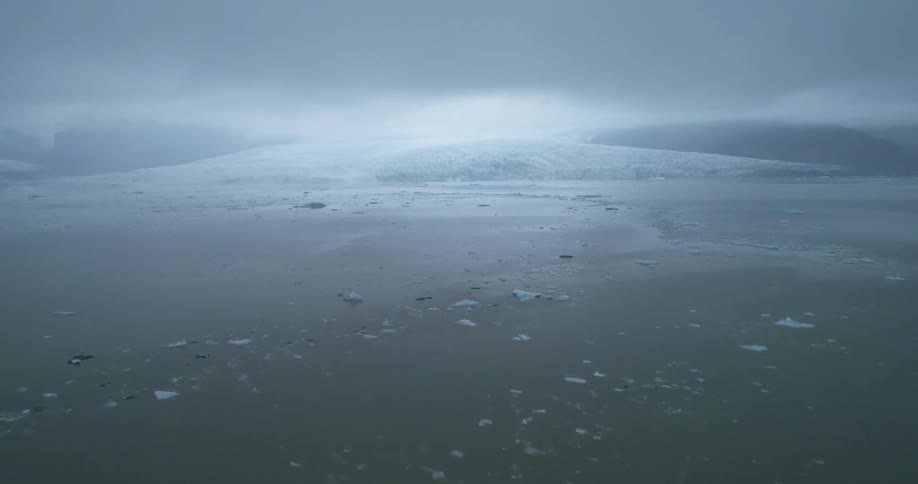 Ice floats on ocean near glacier lit by bright sunlight shining through overcast sky on foggy day