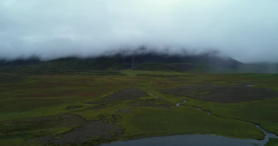 A landscape with grass and water with a mountain range fog the sky and clouds in the background