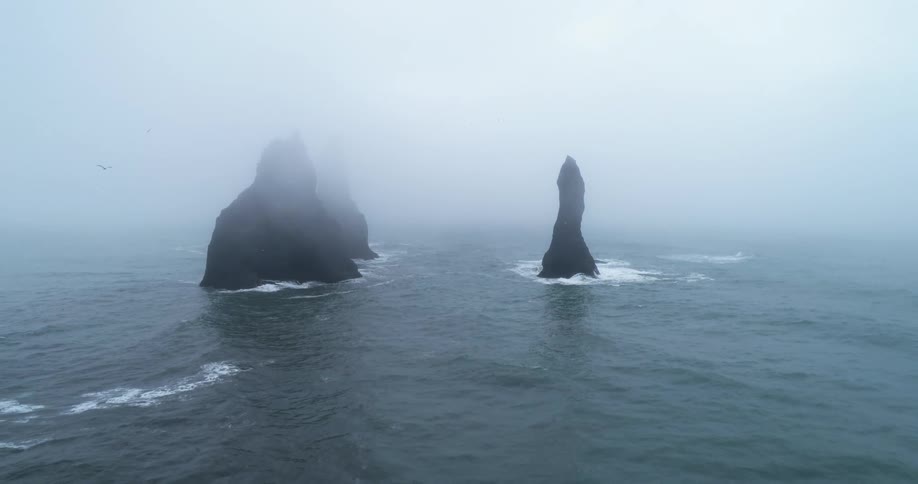 A body of water would rocks jutting out of the water with fog and birds flying in the sky