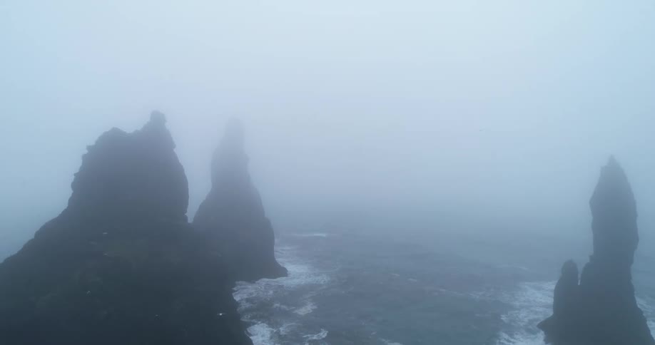 Seagulls fly around tall rock formations in the water on a foggy day