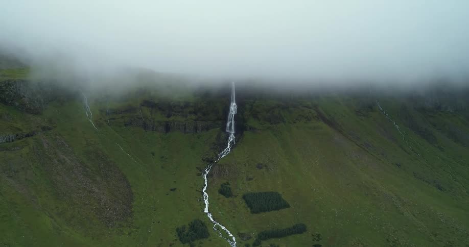 A thick layer of fog hangs over a mountain ridge where a waterfall flows and streams down the green slope