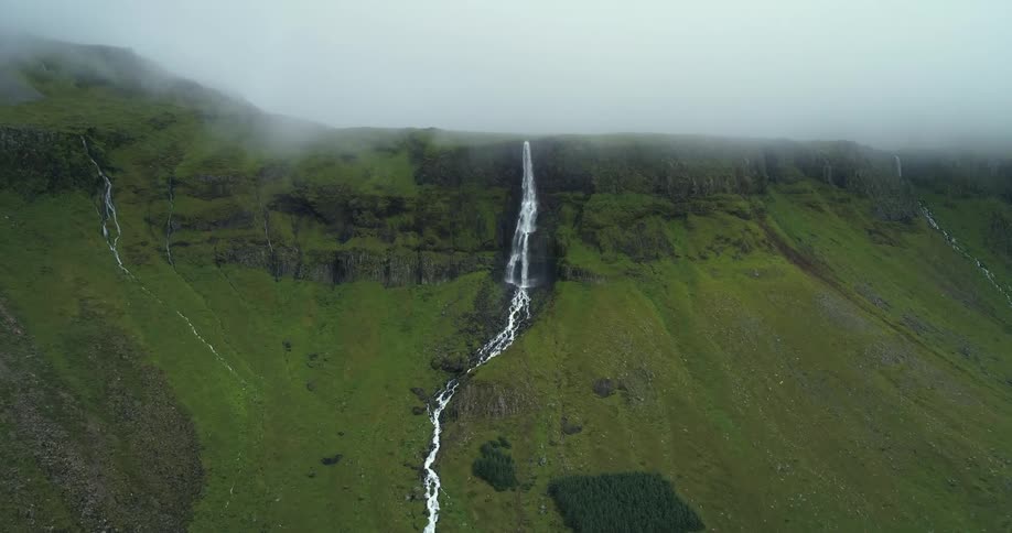 Waterfalls and a stream run down the side of a grass covered hill under a hazy sky