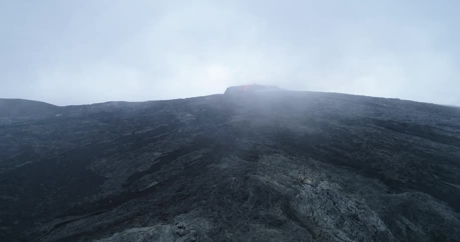 Lava splashes and spills from the top of a volcano under a cloudy sky