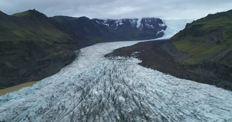An aerial view of a glacier slowly carving its way through a mountain region on a cloudy day