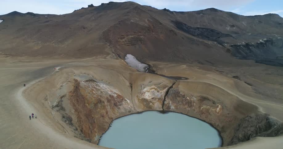 People walking by a crater with a mountain range the sky and clouds in the background