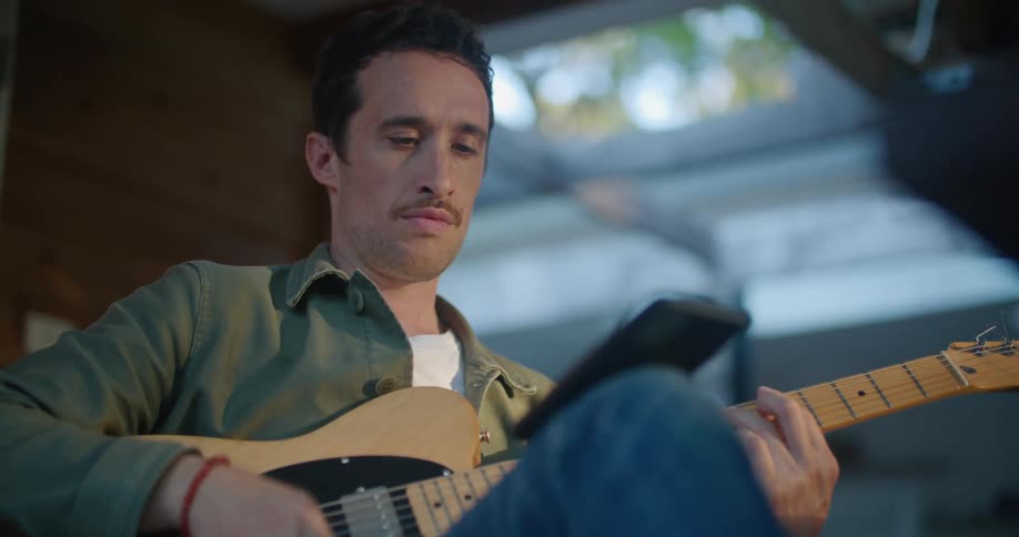 A man sits at home and looks at his cell phone as he plays the guitar