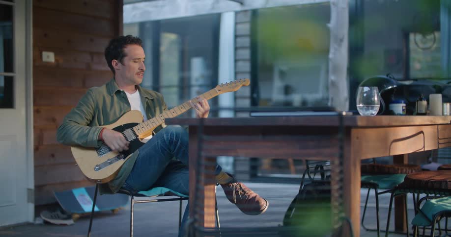 Man sits on porch at table near door playing acoustic guitar