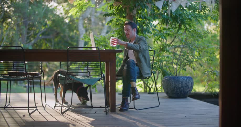 Man sits outdoors in front of laptop with dog playing with a ball nearby