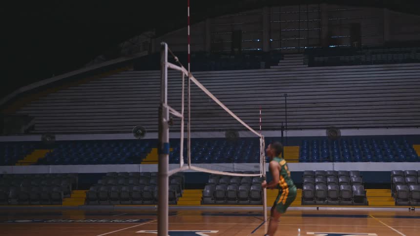 A young man in a jersey is spiking a volleyball over a net inside a gymnasium