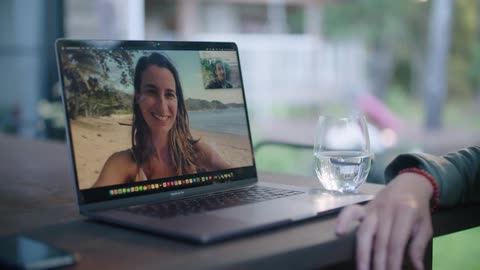 Man sitting at table in backyard talks with woman on beach via laptop they wave and kisses at each other