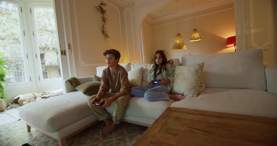 A sister and brother play a video game together while sitting on a sofa in the living room of a house during the day