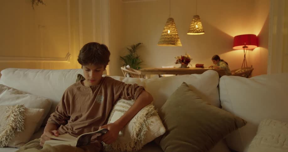 Boy sitting in the couch reading a book in front of a girl sitting on a table doing homework