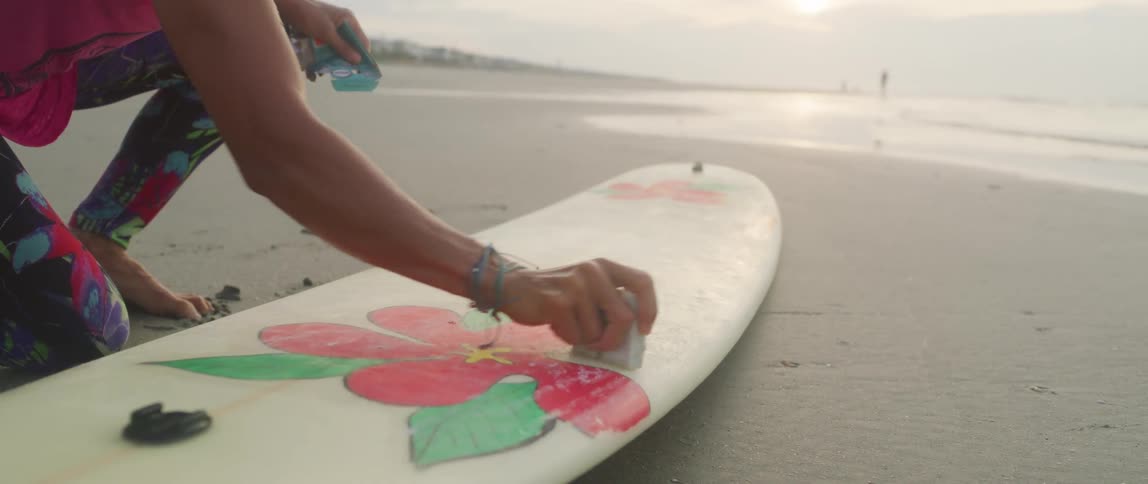 A woman waxes her surfboard while kneeling on the beach