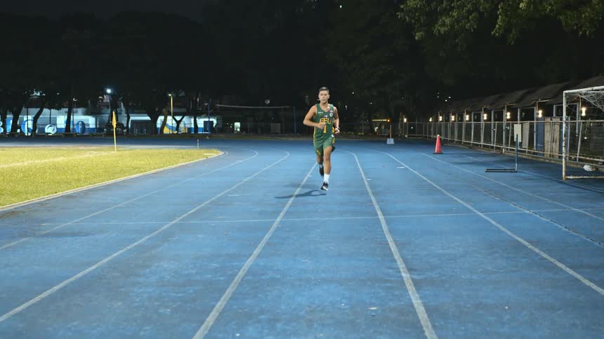 Male athlete in uniform runs along track in athletic field with buildings in background
