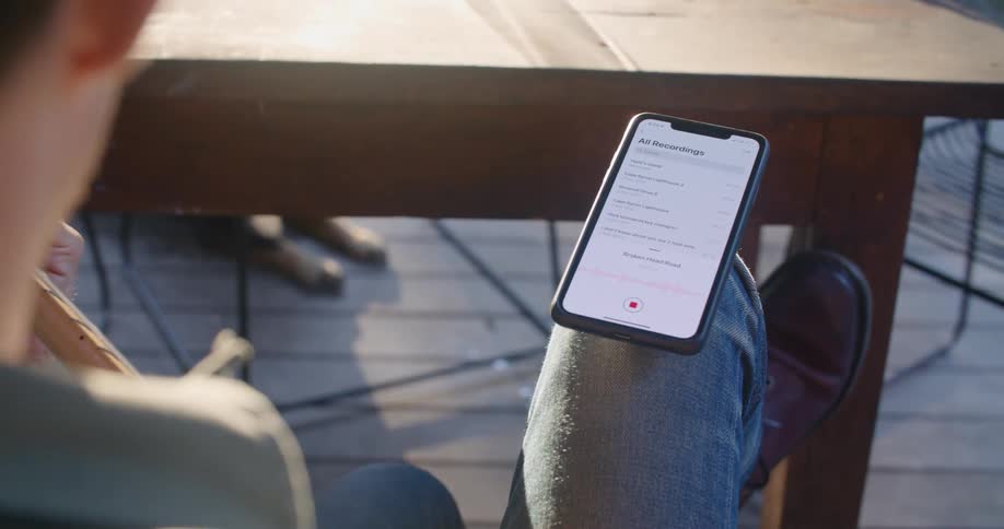 A man is sitting in a chair on a deck strumming a guitar while looking at his cell phone