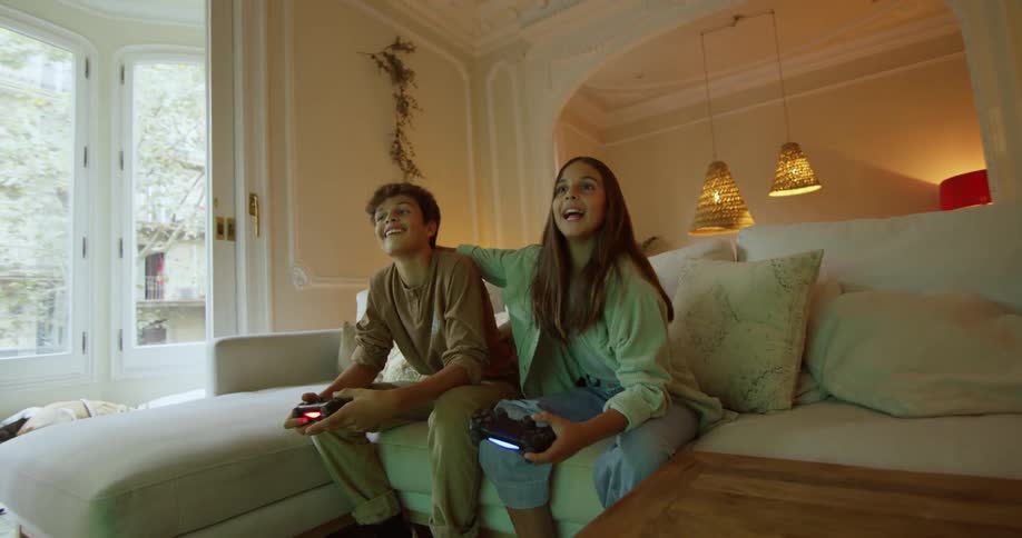 A young boy and girl sit on a couch in a home and laugh as they play video games and the girl covers the boys eyes and tries to distract him