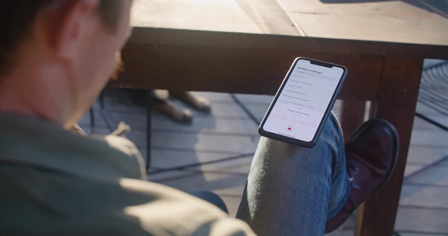 A man is sitting outdoors on a deck recording himself playing a guitar with a cell phone