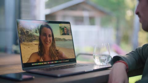 Man with green jacket and earring sits at table and makes a video call with a woman with brown wavy hair on the beach outdoors
