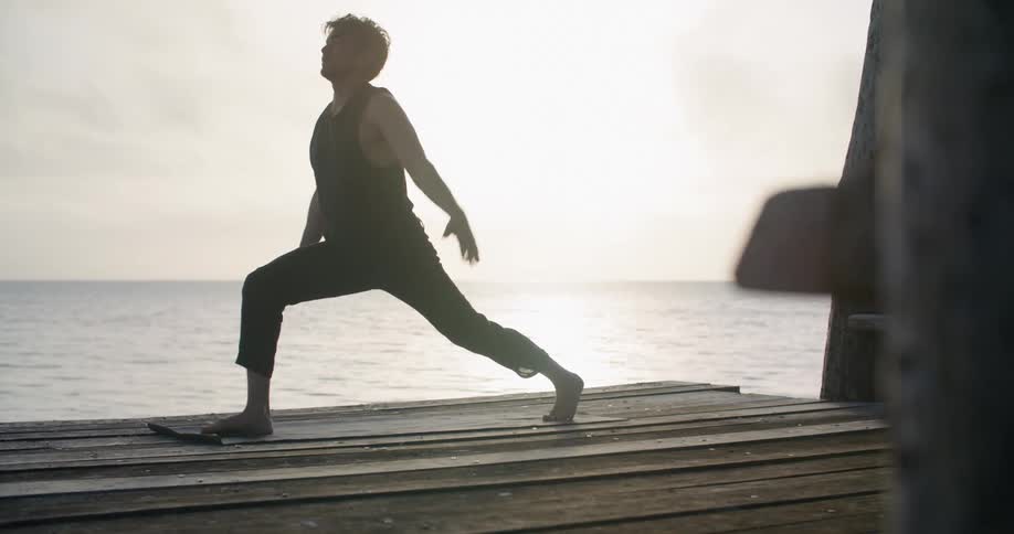 A man kneels and stretches while doing yoga on a pier