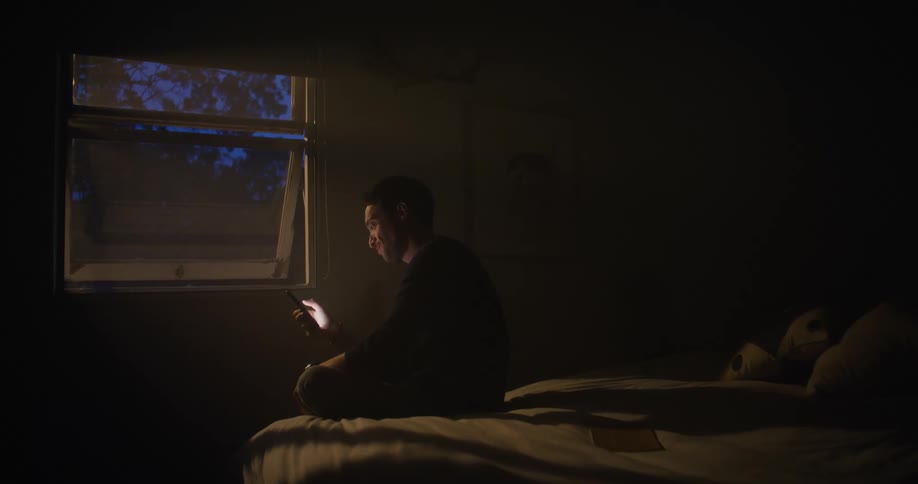 A man is sitting on a bed near an open window and tapping on a cell phone