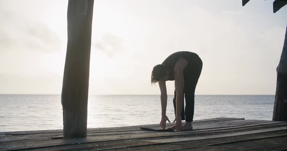 A man is outdoor doing yoga stretches on a wooden deck overlooking the ocean