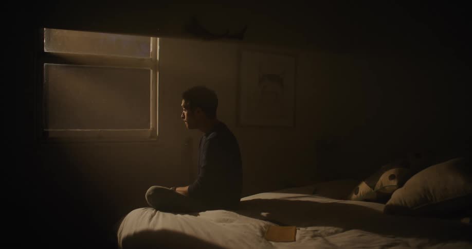 A man is siting on a dark bed while looking out of a window with amber light pouring into the room