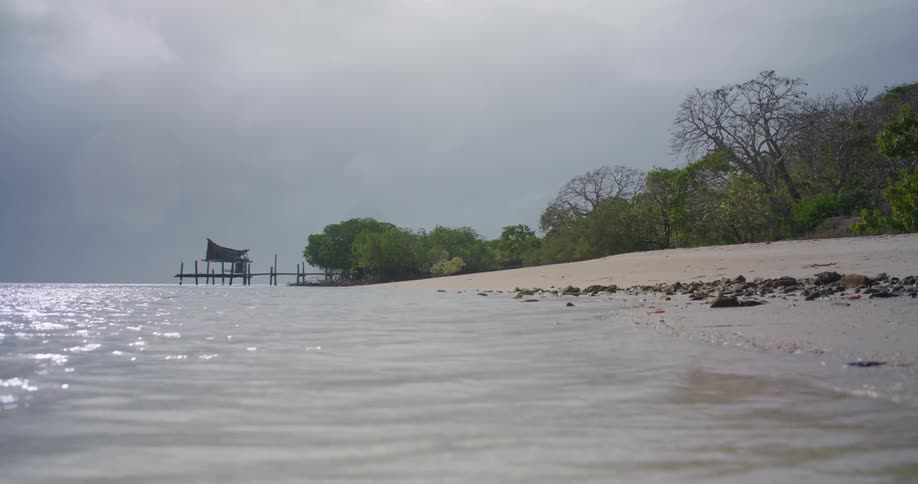 Small waves are flowing onto a beach near an old dock on a cloudy day