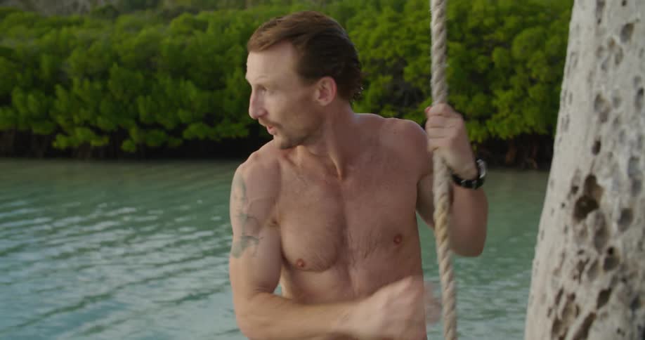A man climbs up a rope onto a wooden platform overlooking a lake surrounded by trees