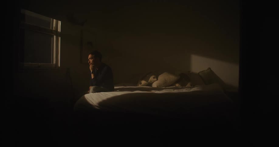 Man with black shirt and jeans sits on bed and looks out the window in dark room