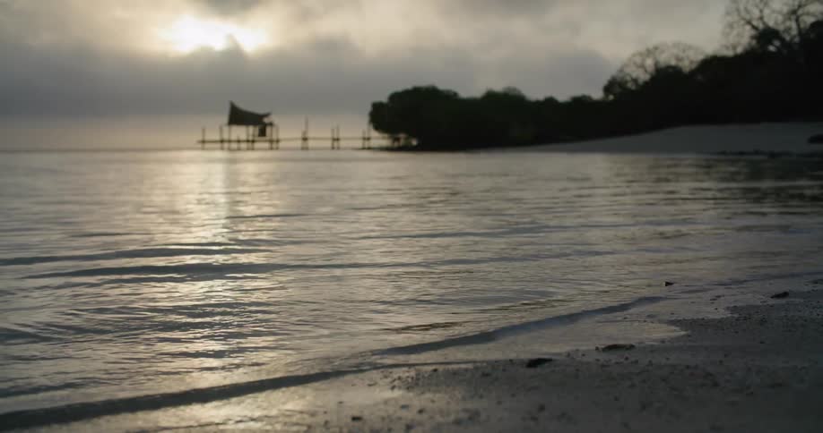 Small waves are gently rolling in onto a beach as sun sets on a cloudy evening