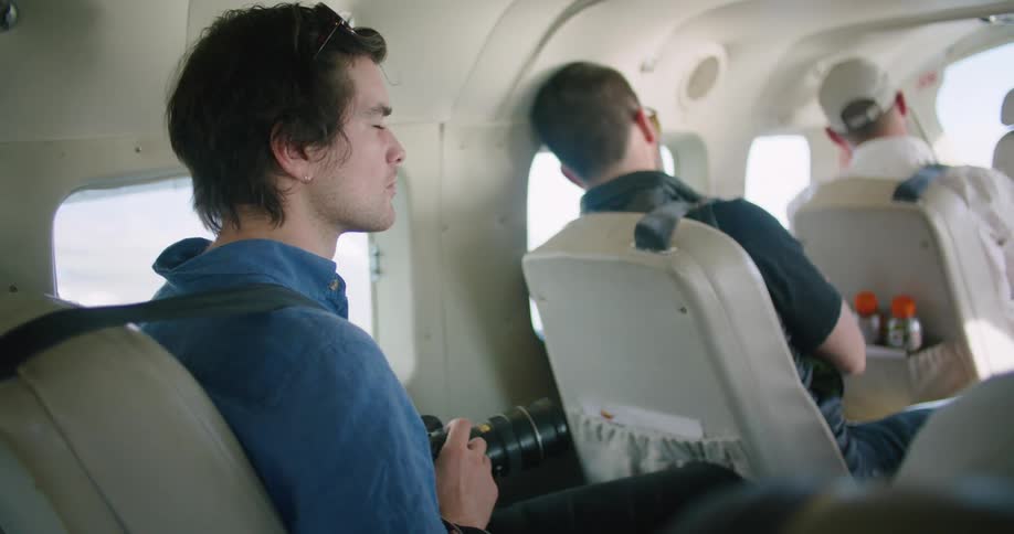 A man holds a camera and looks around while sitting in a small airplane with other passengers
