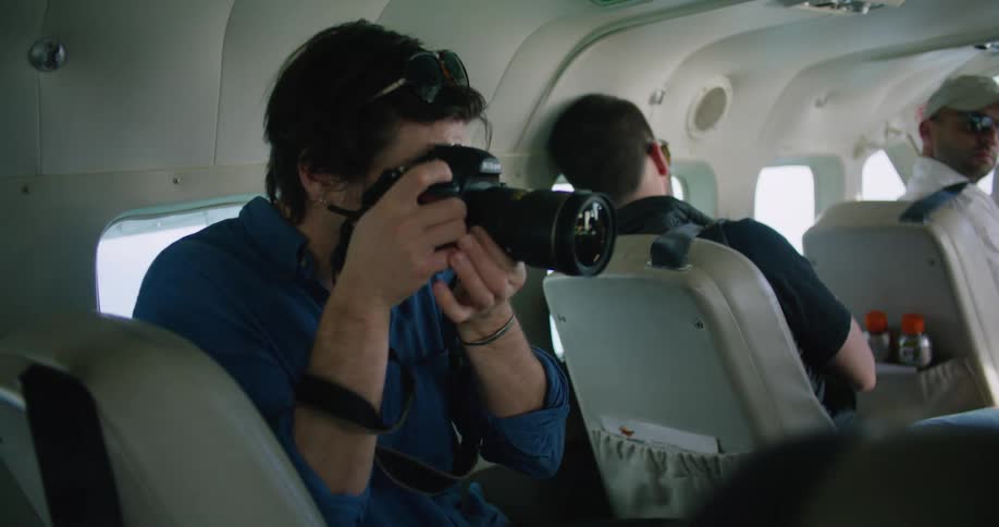 A man sits in the back of a van and takes photos of a person as the driver looks back