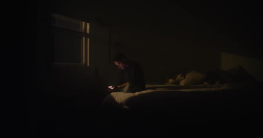 A man sits on his bed at night using his cell phone