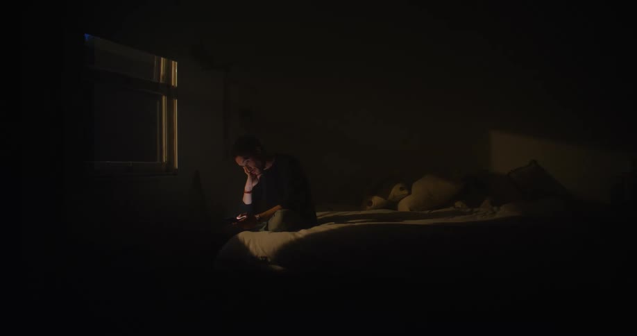 Man sitting on the bed in a dark bedroom looking at his phoe