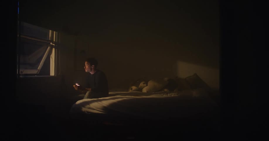 A man texts while sitting at the end of his bed in a dark room