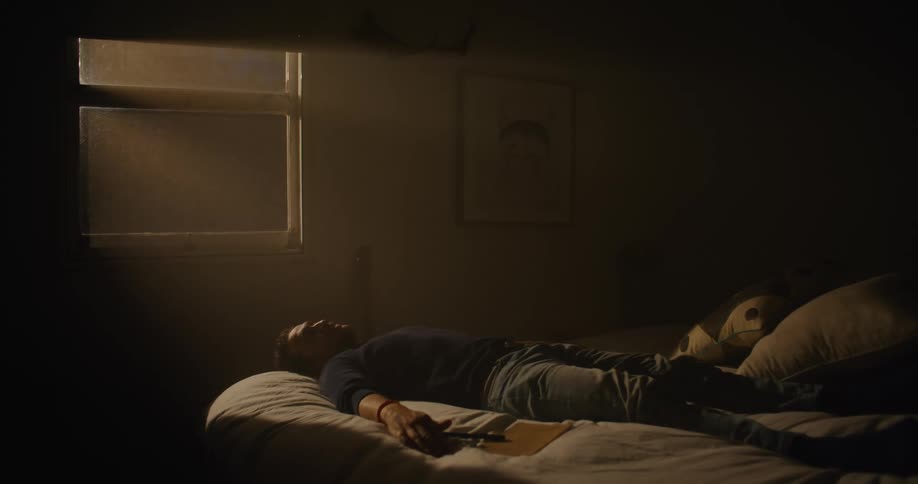 A man is laying on a bed next to a notebook then gets up and walks away