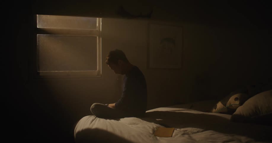 A man is sitting in his dark bedroom looking out the window