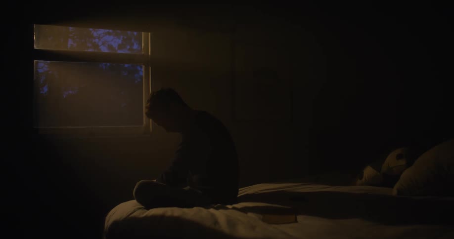 A man is sitting on a bed and staring out a window while putting his head in his hand