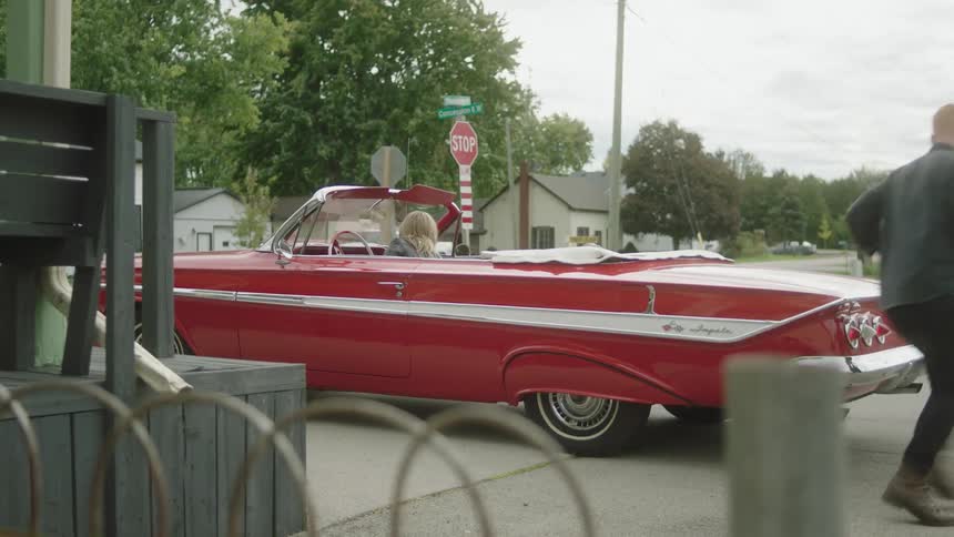 A woman driving a red vintage convertible car is picking up a man with a guitar and duffle bag